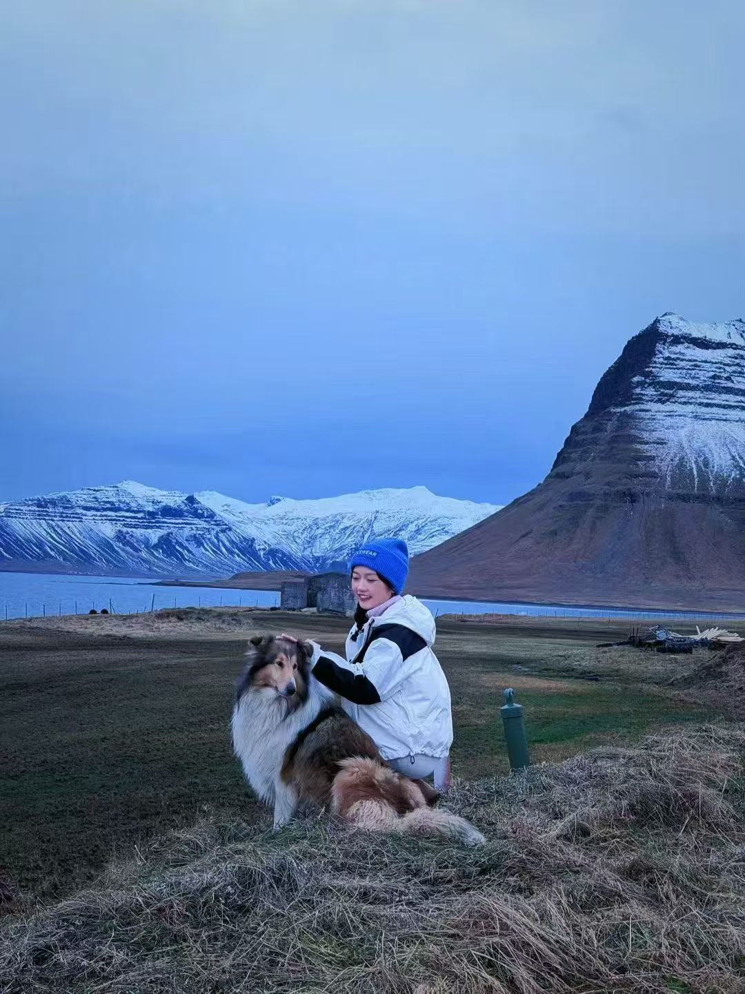女生穿白色外套和蓝帽子在雪山前与狗互动